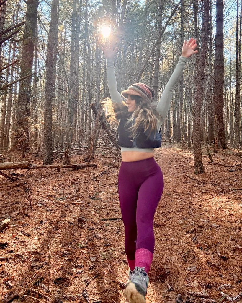 Lauren standing in a sunlit forest with her arms raised