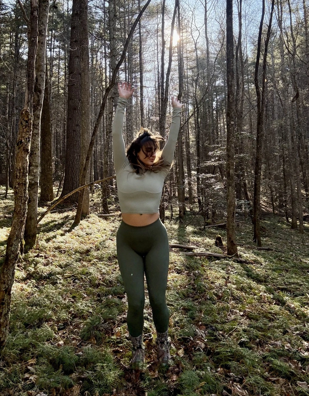 Lauren standing in a sunlit forest with her arms raised to the sky
